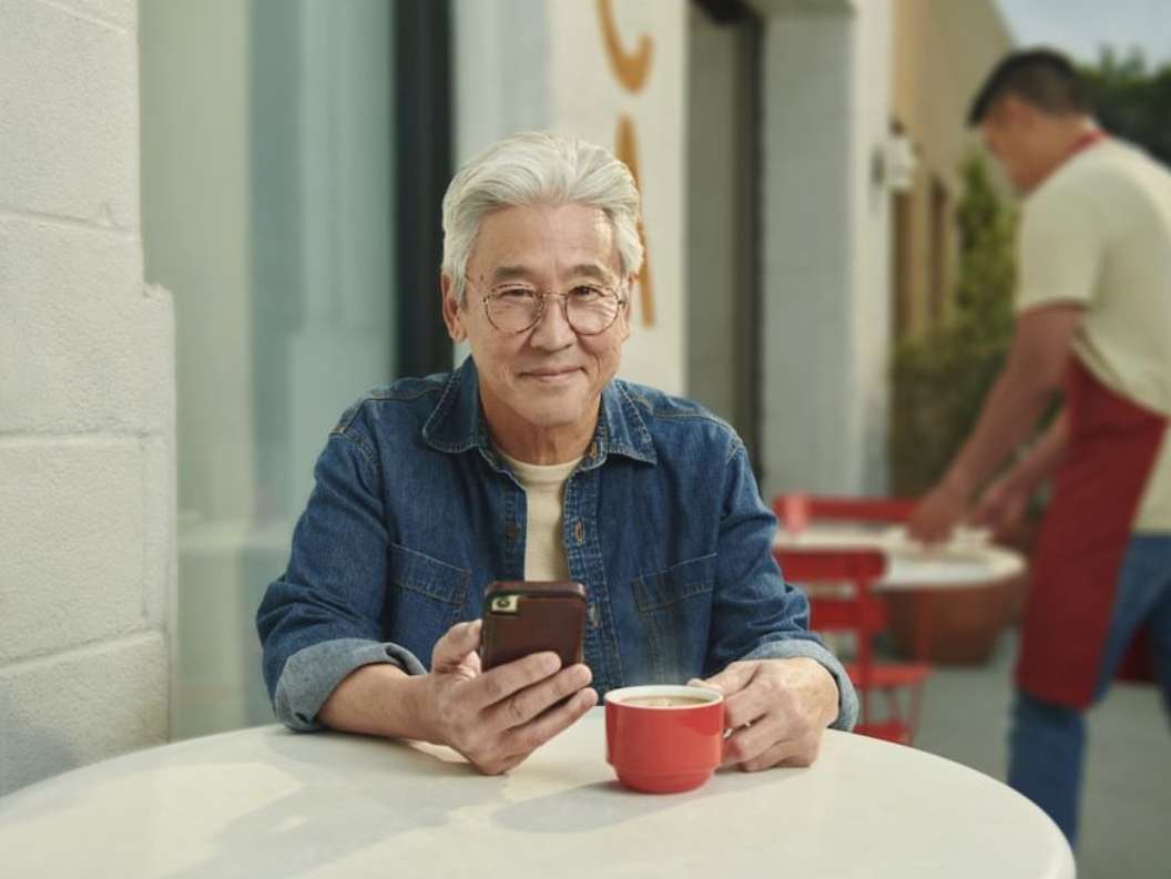 Elderly Asian man smiles while holding his smartphone and sits at a cafe table with a red mug and a waiter in the background.